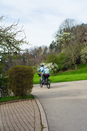 A couple pedals together on a well-maintained path, basking in the beauty of spring flowers and vibrant plants in a peaceful park setting.の写真素材