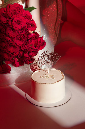 A delicious-looking birthday cake sits next to a vibrant bouquet of red roses. The scene is beautifully lit, creating a festive and romantic atmosphere.の写真素材