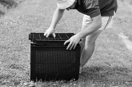 A man carefully places a grid insert into a dark-colored wicker storage container. He's outdoors on grass, assembling the piece.の写真素材