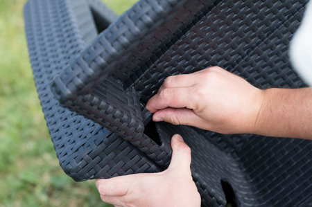 Close-up of hands assembling dark-colored woven plastic outdoor furniture. The person is connecting two parts of the structure.の写真素材