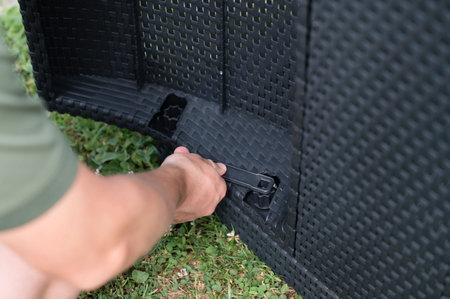 Close-up of a person assembling dark-colored wicker outdoor furniture. They're using a small tool to fasten a section. Green grass is visible in the background.の写真素材