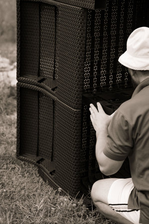 A person assembles dark brown outdoor furniture pieces. The furniture has a woven texture and appears to be made of plastic. The setting is outdoors on grass.の写真素材