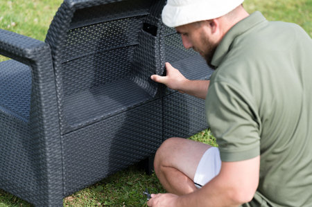 A man assembles dark gray wicker outdoor furniture on a grassy lawn. The piece appears to have storage space. Relax and enjoy the outdoors.の写真素材