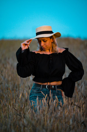 A blonde woman in a black top and jeans stands in a field of wheat. The setting sun casts a warm glow. She gently adjusts her straw hat.の写真素材