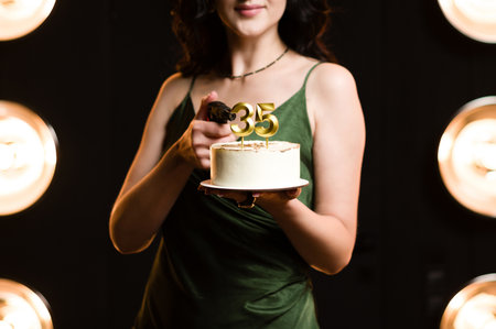 A woman, dressed in a dark green gown, presents a small cake decorated with '35' candles. She holds a handgun in her other hand. The backdrop is a dark room with illuminated round lights.の写真素材