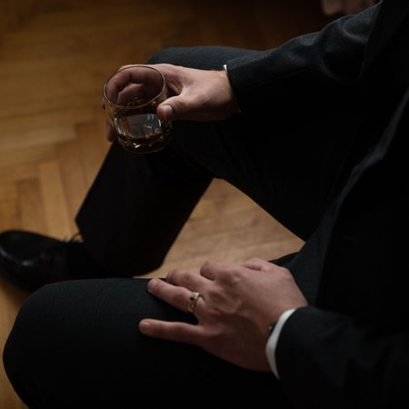 A man in a suit sits and holds a glass of amber liquid, lost in thought.の写真素材