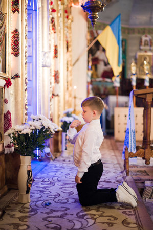 A young boy, dressed in formal attire, kneels in prayer before a floral arrangement within a church setting. The scene is peaceful and contemplative.の写真素材