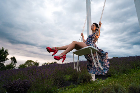 A carefree woman in a flowing dress swings high, her red heels adding a pop of color against the...