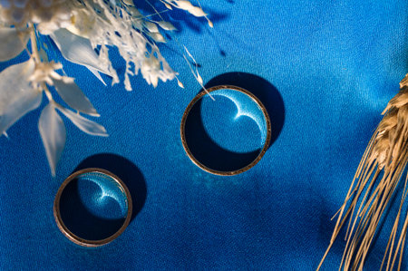 Close-up shot of two gold wedding bands resting on a deep blue background. White flowers and wheat stalks add a touch of elegance and rustic charm.の写真素材