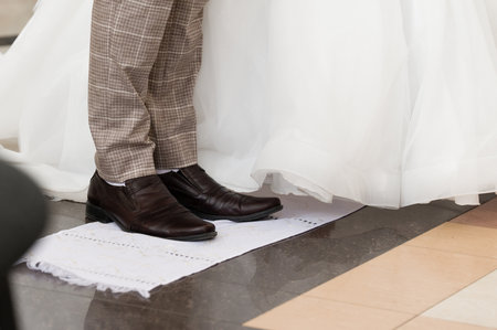 Close-up of a groom's feet in brown leather shoes and patterned pants. Part of a white wedding dress is visible in the background. The scene suggests a wedding ceremony.の写真素材