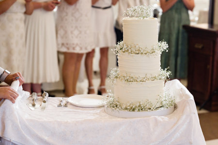 A pristine white wedding cake, three tiers high, decorated with small white flowers sits on a table. Guests in white dresses are blurred in the background.の写真素材