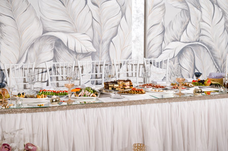A beautifully set wedding reception table features an array of appetizers, cakes, and desserts. White chairs and a stylish backdrop enhance the elegant atmosphere.の写真素材