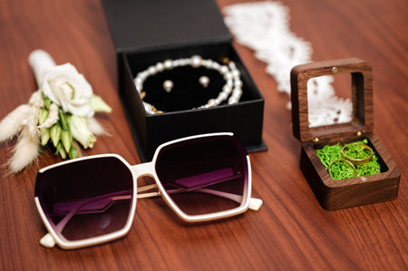 A stylish arrangement of wedding essentials: elegant sunglasses, pearl jewelry in a box, and wedding rings nestled in a wooden case. The scene is set on a rich wooden surface.の写真素材