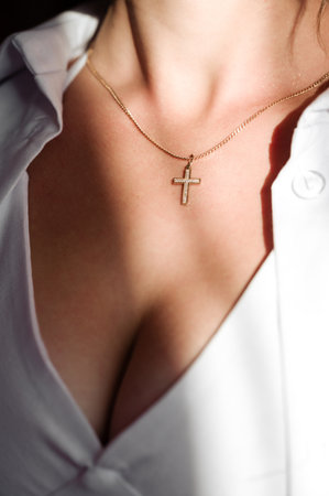 A woman's decolletage is shown, adorned with a delicate gold chain featuring a small, sparkling cross pendant. The image emphasizes the detail of the jewelry against the soft skin.の写真素材
