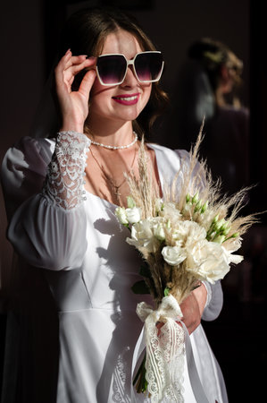 A radiant bride in a simple white dress poses, holding a bouquet of white flowers and pampas grass. She wears stylish sunglasses, adding a touch of modern flair to her classic bridal look.の写真素材