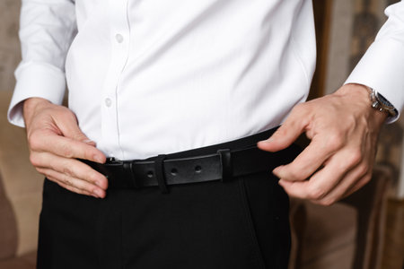 Close-up view of a person fastening a black leather belt over dark trousers and a crisp white shirt. The image focuses on the details of the clothing and accessories.の写真素材