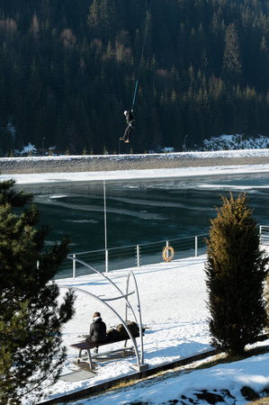 A winter day brings excitement to life as a person ziplines over a still frozen lake, surrounded by snow-covered slopes and lush green conifers in the distance.の写真素材