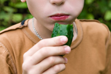 Close-up of a child enjoying a fresh cucumber outside. The focus is on the child's mouth and hand holding the partially eaten cucumber, suggesting a healthy snack or mealtime.の写真素材