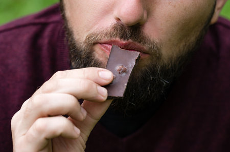 Close-up of a bearded man savoring a piece of dark chocolate. The focus is on the chocolate and his...