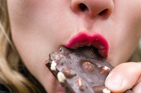 Close-up of a woman with pink lipstick taking a bite of a dark chocolate bar with nuts. The focus is...