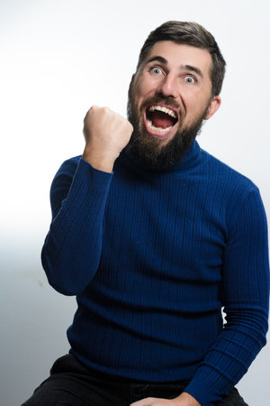 A bearded man in a blue sweater and dark pants sits against a white background, expressing joy with a raised fist and open mouth.の写真素材
