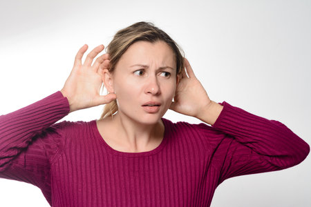 A woman with a concerned expression cups her hands around her ears, attempting to improve her...