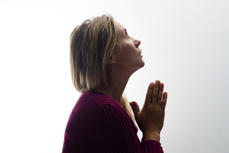 Side profile of a woman with clasped hands, her head tilted up in prayer against a bright...