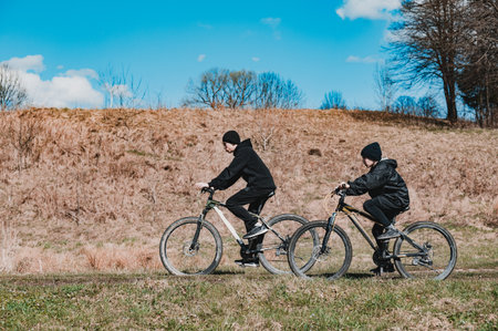 Two people on mountain bikes ride along a path. The setting is a grassy field with a hill in the...
