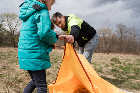 In a beautiful spring landscape, a father and daughter work together, carefully assembling an orange tent as dark clouds loom overhead. Their excitement is palpable.の写真素材