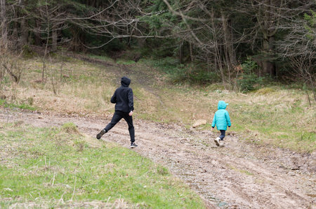 A child and an adult navigate a muddy path through a serene forest, each dressed for cool weather....