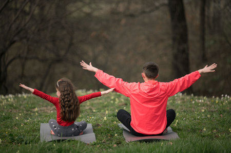In a quiet forest, a child and an adult enjoy a moment of harmony while practicing yoga. They stretch their arms wide, absorbed in the calm of nature on a sunny day.の写真素材