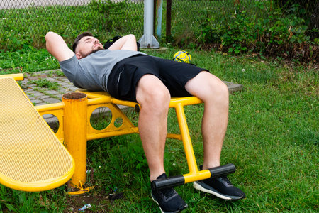 A man performs sit-ups on yellow outdoor gym equipment. He's wearing a gray shirt and black shorts. The setting is a grassy park.の写真素材