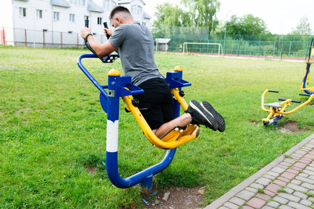 A man uses an outdoor workout machine. The setting is a park, with green grass and a building visible. He appears focused on his fitness routine.の写真素材