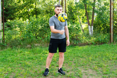 A man with a beard juggles a soccer ball in a grassy park. He's wearing a grey shirt and black shorts. A relaxed, sunny day.の写真素材