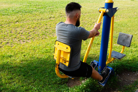 A man uses an outdoor exercise machine in a grassy park setting. The machine is yellow and blue....