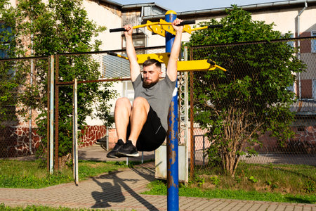 A man performs hanging leg raises on outdoor exercise equipment. He's focused and strong,...