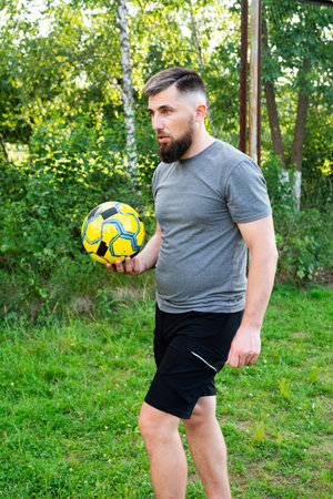 A bearded man in athletic attire holds a soccer ball. He's standing on a grassy field, possibly a park or recreational area. The setting is outdoors and natural.の写真素材
