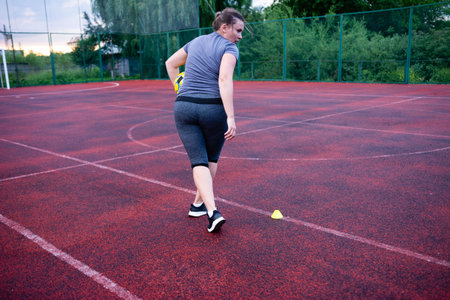 A female athlete performs soccer training exercises on a red outdoor court. She holds a ball and approaches a training cone. The scene is focused on her athletic activity.の写真素材