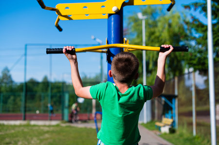 A young boy performs pull-ups on an outdoor exercise machine. The sunny day and green surroundings create a positive and active atmosphere.の写真素材