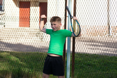 A playful image of a boy in a green shirt and black shorts, holding a tennis racquet, posing confidently near a chain-link fence on a sunny day.の写真素材
