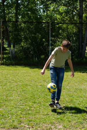 A young boy skillfully juggles a soccer ball outdoors. He's on a grassy field, near a fence, enjoying a sunny day. The scene is peaceful and captures a moment of playful activity.の写真素材