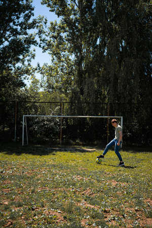 A teenager enjoys a sunny afternoon, practicing his soccer skills on an outdoor field. He kicks a ball towards a goal, surrounded by lush greenery and trees.の写真素材