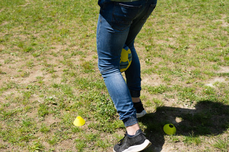 Close-up view of a person's legs in jeans, holding a soccer ball. They appear to be performing drills on a grassy field with yellow training cones nearby. Sunny outdoor setting.の写真素材