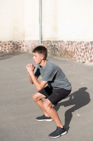 A young man performs squats as part of his outdoor fitness routine. He's concentrating on his form against a stone wall backdrop. The sunlit scene conveys energy and health.の写真素材