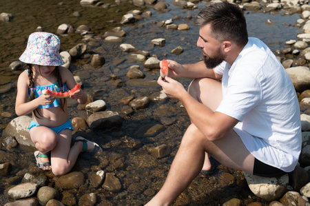 A man and young girl, both in summer attire, sit amongst smooth river rocks, happily eating watermelon slices. The shallow, clear water flows gently around them.の写真素材