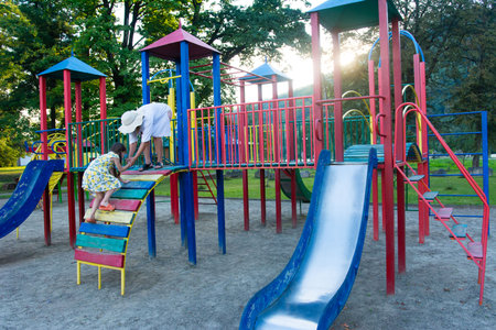 A young girl and an adult are on a brightly colored playground. The girl is climbing a wooden ramp...