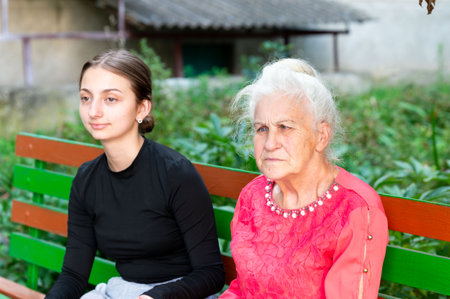 A young woman and an elderly lady sit side by side on a vibrant green bench, surrounded by verdant foliage, enjoying a peaceful moment together in the late afternoon light.の写真素材