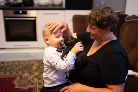 A grandmother smiles warmly while her baby grandson, with a pacifier, pets their black cat. The...