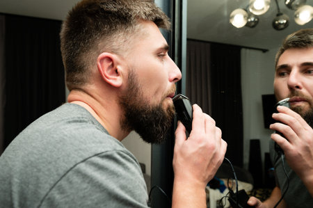 In a contemporary living space, a man skillfully uses an electric trimmer to groom his beard while closely examining his reflection in a sleek mirror.の写真素材