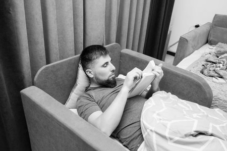 A man comfortably nestled on a couch reads intently, surrounded by soft pillows and blankets, enveloped in a serene atmosphere perfect for unwinding after a long day.の写真素材